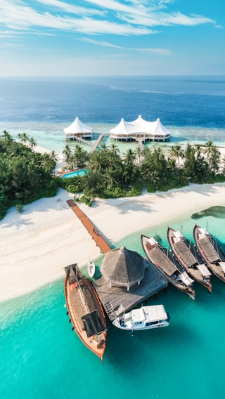 A tropical island features pristine white sandy beaches, lush green palm trees, and turquoise blue waters. A group of traditional wooden boats and a small yacht are docked at a wooden pier. In the background, a luxury resort with white tent-like structures is visible on the water.