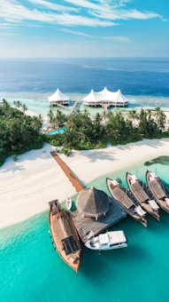A tropical island features pristine white sandy beaches, lush green palm trees, and turquoise blue waters. A group of traditional wooden boats and a small yacht are docked at a wooden pier. In the background, a luxury resort with white tent-like structures is visible on the water.