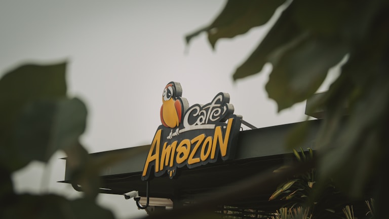 A colorful sign featuring a cartoon parrot representing a cafe named Amazon, partially obscured by green leaves in the foreground.