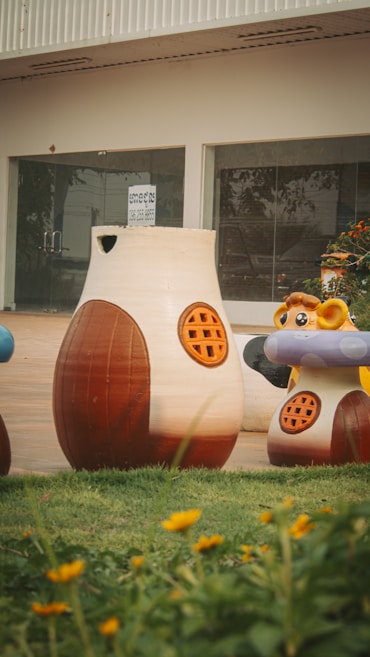 Large animal-themed play structures resembling a bear and a giraffe stand on a grassy patch, set against a background of a glass-fronted building. Brightly colored flowers bloom in the foreground.