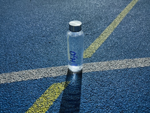 A clear plastic water bottle labeled H2O stands upright on a textured blue surface, intersected by white and yellow lines, likely part of a sports track.