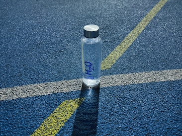 A clear plastic water bottle labeled H2O stands upright on a textured blue surface, intersected by white and yellow lines, likely part of a sports track.