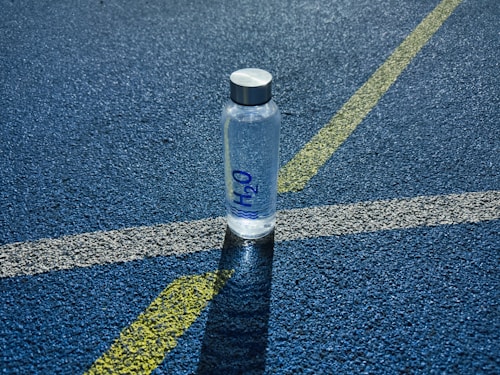 A clear plastic water bottle labeled H2O stands upright on a textured blue surface, intersected by white and yellow lines, likely part of a sports track.