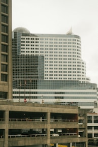 A modern multi-story office building with a white and glass facade, including a domed section at the top. Multiple flags are visible on the lower part of the building. In the foreground, there is a concrete parking structure with multiple levels, including visible vehicles.