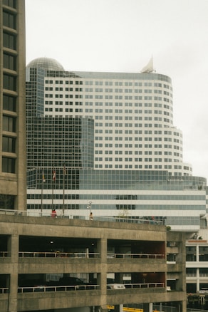 A modern multi-story office building with a white and glass facade, including a domed section at the top. Multiple flags are visible on the lower part of the building. In the foreground, there is a concrete parking structure with multiple levels, including visible vehicles.