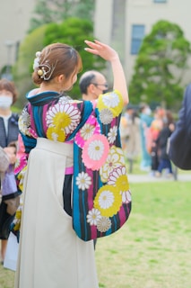 A person wearing a colorful kimono with floral patterns stands outdoors in a grassy area. The kimono features vibrant colors such as pink, yellow, and blue with large flower designs. The individual's hair is neatly styled with decorative hairpieces. A crowd of other people can be seen in the background, some wearing masks and traditional clothing.