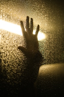 A silhouetted hand is pressed against a rain-speckled glass surface, with light shining from behind, creating a dramatic backlight effect. The image conveys a sense of intimacy and mystery.