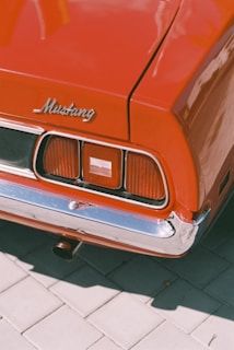 Close-up of the rear section of a vintage red Mustang car showcasing the Mustang emblem, tail lights, and chrome bumper against a tiled pavement.