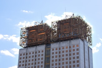 A modern high-rise building features two symmetrical structures on the rooftop, enclosed by intricate scaffolding. The facade is lined with square windows, and the structure is set against a clear blue sky with some white clouds, adding a sense of openness and height.