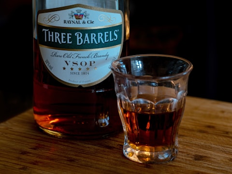 A bottle of Raynal & Cie brandy labeled 'Three Barrels' with the designation VSOP sits on a wooden surface. Next to it is a glass partially filled with the amber liquid.