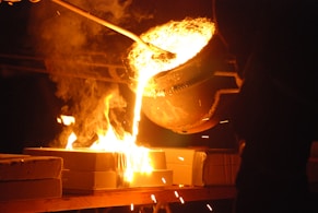Molten metal is being poured from a large ladle into molds, creating bright orange and yellow flames. Sparks fly as the glowing liquid metal cascades downward, illuminating the surrounding area with intense light.
