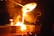 Molten metal is being poured from a large ladle into molds, creating bright orange and yellow flames. Sparks fly as the glowing liquid metal cascades downward, illuminating the surrounding area with intense light.