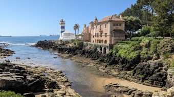 A coastal scene features a charming, large building with multiple windows and an intricate design, positioned near the edge of rocky terrain. A lighthouse with red and white stripes stands nearby, overlooking the ocean. There is a winding, narrow waterway leading from the ocean into the rocky shore, bordered by lush greenery and trees on the right side.