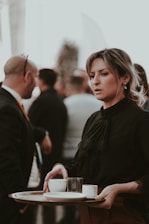 A woman wearing a black blouse is holding a tray with cups at what appears to be a social gathering or formal event. She has her hair tied back, and several people can be seen in the background, some with blurred details.
