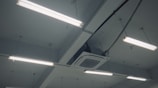 Fluorescent lights are mounted on the ceiling along with an air conditioning unit. The ceiling is structured with beams, and several cables and wires are connected to the lights and the unit.
