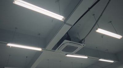 Fluorescent lights are mounted on the ceiling along with an air conditioning unit. The ceiling is structured with beams, and several cables and wires are connected to the lights and the unit.