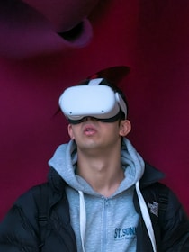 A person wearing a virtual reality headset appears to be engaged or immersed in the experience. They are dressed in a casual outfit consisting of a hoodie and a jacket, with a light-colored background offering a contrast to the darker tones of the clothing.