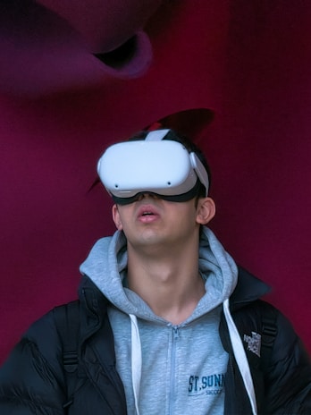 A person wearing a virtual reality headset appears to be engaged or immersed in the experience. They are dressed in a casual outfit consisting of a hoodie and a jacket, with a light-colored background offering a contrast to the darker tones of the clothing.