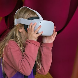 A young girl with long blonde hair is using a white virtual reality headset. She is wearing a long-sleeved, pinkish-red shirt, and appears to be engaged and curious. The background features deep pink and purple colors in a soft, abstract pattern.