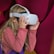 A young girl with long blonde hair is using a white virtual reality headset. She is wearing a long-sleeved, pinkish-red shirt, and appears to be engaged and curious. The background features deep pink and purple colors in a soft, abstract pattern.