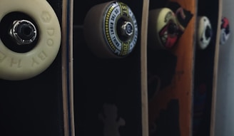 A close-up view of several skateboard decks aligned vertically, each attached with different wheels. Some decks have unique designs and graphics, with visible hardware like bolts and bearings. The lighting creates a dramatic effect with shadows and highlights on the textured surfaces.
