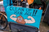 A humorous street sign featuring a cartoon illustration of a poop emoji flexing muscular arms on a blue background, with the words 'Clean Up Your Act!!'. The sign is attached to a bicycle, and the scene appears to be set on a rainy day in an urban environment.