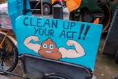 A humorous street sign featuring a cartoon illustration of a poop emoji flexing muscular arms on a blue background, with the words 'Clean Up Your Act!!'. The sign is attached to a bicycle, and the scene appears to be set on a rainy day in an urban environment.