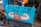 A humorous street sign featuring a cartoon illustration of a poop emoji flexing muscular arms on a blue background, with the words 'Clean Up Your Act!!'. The sign is attached to a bicycle, and the scene appears to be set on a rainy day in an urban environment.