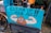 A humorous street sign featuring a cartoon illustration of a poop emoji flexing muscular arms on a blue background, with the words 'Clean Up Your Act!!'. The sign is attached to a bicycle, and the scene appears to be set on a rainy day in an urban environment.
