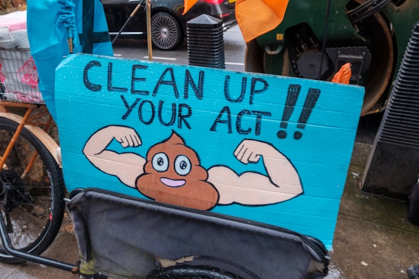 A humorous street sign featuring a cartoon illustration of a poop emoji flexing muscular arms on a blue background, with the words 'Clean Up Your Act!!'. The sign is attached to a bicycle, and the scene appears to be set on a rainy day in an urban environment.