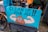 A humorous street sign featuring a cartoon illustration of a poop emoji flexing muscular arms on a blue background, with the words 'Clean Up Your Act!!'. The sign is attached to a bicycle, and the scene appears to be set on a rainy day in an urban environment.