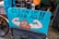 A humorous street sign featuring a cartoon illustration of a poop emoji flexing muscular arms on a blue background, with the words 'Clean Up Your Act!!'. The sign is attached to a bicycle, and the scene appears to be set on a rainy day in an urban environment.