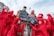 A group of people wearing red costumes and face paint surround two individuals holding signs. The signs contain environmental messages, emphasizing the conflict between planet preservation and profit. The scene captures a protest or demonstration with participants expressing concern for environmental issues.