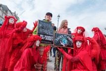 A group of people wearing red costumes and face paint surround two individuals holding signs. The signs contain environmental messages, emphasizing the conflict between planet preservation and profit. The scene captures a protest or demonstration with participants expressing concern for environmental issues.