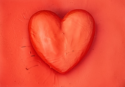 A three-dimensional red heart shape sits against a textured red background, with subtle shadows and highlights creating depth and dimension.