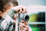 A person is holding a silver camera close to their face, capturing a photo. The camera lens is prominently visible, and the person's hand is on the camera. The background is blurred, focusing the attention on the camera and the person.