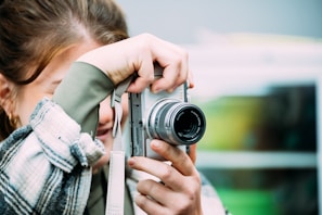 A person is holding a silver camera close to their face, capturing a photo. The camera lens is prominently visible, and the person's hand is on the camera. The background is blurred, focusing the attention on the camera and the person.