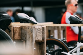 A blurred background features people, with the focus on a bicycle rack made of wooden pallets labeled 'EPAL'. Bicycle seats and parts are visible, suggesting a bike storage or repair area. One person is wearing a red and white cycling jersey and sunglasses.