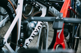 A close-up view of several high-performance racing bicycles oriented tightly together. The image focuses on the frames and wheels, showcasing brand names and sleek designs. There are vibrant colors and elements such as water bottles and intricate handlebar setups.