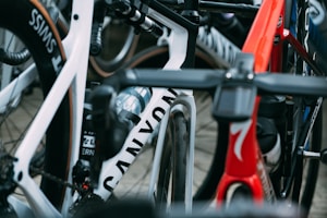 A close-up view of several high-performance racing bicycles oriented tightly together. The image focuses on the frames and wheels, showcasing brand names and sleek designs. There are vibrant colors and elements such as water bottles and intricate handlebar setups.