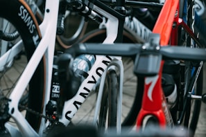 A close-up view of several high-performance racing bicycles oriented tightly together. The image focuses on the frames and wheels, showcasing brand names and sleek designs. There are vibrant colors and elements such as water bottles and intricate handlebar setups.