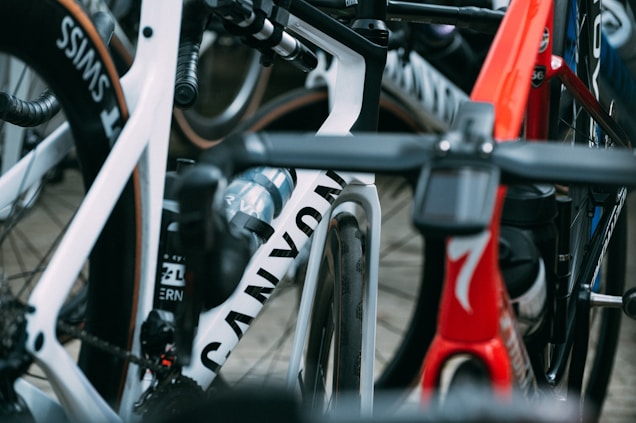 A close-up view of several high-performance racing bicycles oriented tightly together. The image focuses on the frames and wheels, showcasing brand names and sleek designs. There are vibrant colors and elements such as water bottles and intricate handlebar setups.