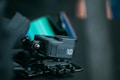 A close-up of a compact action camera placed on a dark surface. The camera is black with branding visible on the side. There is a hint of vibrant color, possibly from a reflection or another object in the background.