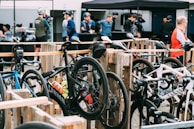 Several road bikes are neatly parked in a designated area made from wooden pallets. Cyclists in riding gear and helmets gather in the background, possibly taking a break or preparing for a cycling event. A tent is set up in the background with more people gathered around what seems to be a refreshment or registration table.
