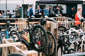Several road bikes are neatly parked in a designated area made from wooden pallets. Cyclists in riding gear and helmets gather in the background, possibly taking a break or preparing for a cycling event. A tent is set up in the background with more people gathered around what seems to be a refreshment or registration table.