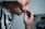 A person is carefully using a straight razor to shave their face, with one hand holding the razor and the other pulling the skin taut. They are wearing a black and white striped shirt and are positioned in front of a mirror which reflects part of their image.