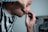 A person is carefully using a straight razor to shave their face, with one hand holding the razor and the other pulling the skin taut. They are wearing a black and white striped shirt and are positioned in front of a mirror which reflects part of their image.