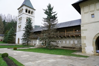A historic building with a tall, white bell tower next to a stone structure featuring wooden beams and a slate roof. Several trees are planted nearby, with well-maintained lawns and a paved pathway leading to the entrance. The surrounding area includes a few plaques on the wall and manicured bushes.