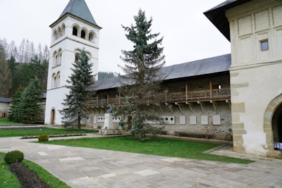 A historic building with a tall, white bell tower next to a stone structure featuring wooden beams and a slate roof. Several trees are planted nearby, with well-maintained lawns and a paved pathway leading to the entrance. The surrounding area includes a few plaques on the wall and manicured bushes.