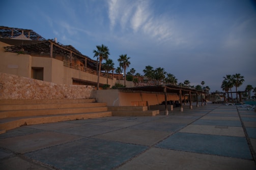 An outdoor area featuring a large stone and concrete tiled patio with steps leading up to a building on the left. The building has a rustic, natural look with exposed stonework and wooden pergolas. Palm trees are scattered throughout the scene, some of which are next to the building. The sky is partially cloudy with a blue and warm golden hue indicating either sunrise or sunset.